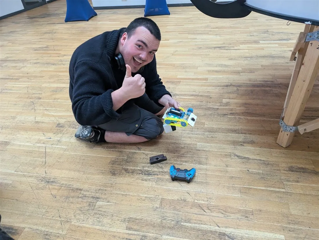 Ein Maker sitzt auf dem Hallenboden, hält einen kleinen Kettenroboter und zeigt den Daumen nach oben